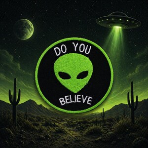 Może przedstawiać: Okrągła czarna naszywka z zieloną twarzą kosmity i białym napisem "DO YOU BELIEVE". Naszywka jest umieszczona na tle pustynnego krajobrazu z UFO i zieloną planetą w tle.
