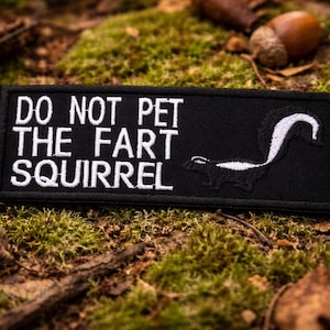 May include: Black rectangular patch with white embroidered text that reads "DO NOT PET THE FART SQUIRREL." A skunk illustration is on the right. The patch is surrounded by moss, leaves, and acorns.