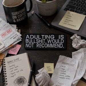 Könnte beinhalten: Eine schwarze Kaffeetasse mit dem Text "SURVIVING ON COFFEE & ANXIETY" steht neben einem Aufnäher mit der Aufschrift "ADULTING IS BULLSHIT. WOULD NOT RECOMMEND." Weitere Artikel sind Rechnungen, ein Laptop mit einer To-Do-Liste und eine Einkaufsliste.
