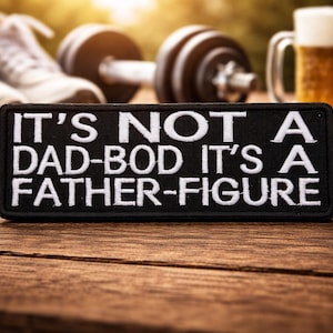 May include: A black patch with white embroidered text that reads "IT'S NOT A DAD-BOD IT'S A FATHER-FIGURE" sits on a wooden surface. In the background are a pair of sneakers, a dumbbell, and a beer.