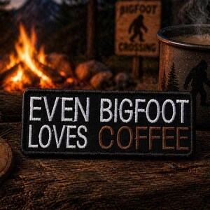 Può includere: Una toppa nera con testo ricamato bianco che recita "EVEN BIGFOOT LOVES COFFEE". Una tazza a tema Bigfoot con caffè è accanto alla toppa. Un cartello di attraversamento di Bigfoot e un falò sono sullo sfondo.
