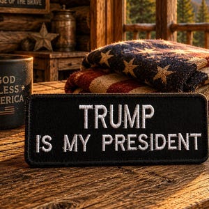 May include: A black mug with "GOD BLESS AMERICA" text and an American flag design sits next to a patch that reads "TRUMP IS MY PRESIDENT". An American flag blanket and a brown cap with an American flag are also visible.