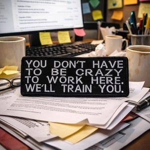 May include: A black rectangular patch with white embroidered text that reads "YOU DON'T HAVE TO BE CRAZY TO WORK HERE. WE'LL TRAIN YOU." The patch is on a desk with papers, coffee mugs, and a computer monitor.