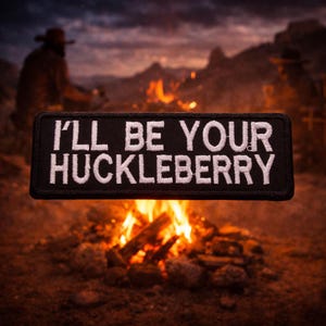 May include: Black rectangular patch with white embroidered text that reads "I'LL BE YOUR HUCKLEBERRY." The patch is set against a blurred background of a campfire and a person in a cowboy hat.