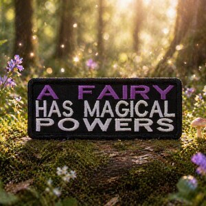 Puede incluir: Un parche rectangular negro con el texto "A FAIRY HAS MAGICAL POWERS" en letras moradas y blancas. El parche está situado en una escena de bosque con flores y setas.