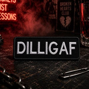 May include: A black rectangular patch with the white embroidered text "DILLIGAF". The patch is on a dark, textured surface with a silver chain and safety pin. A glass of amber liquid is visible in the background.