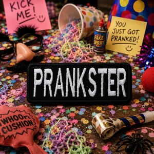 Puede incluir: Un parche rectangular negro con la palabra "PRANKSTER" bordada en blanco, rodeado de artículos de fiesta. Una nota adhesiva "Kick Me!", un "Whoopee Cushion" y una lata de "Fake Spray" también son visibles.