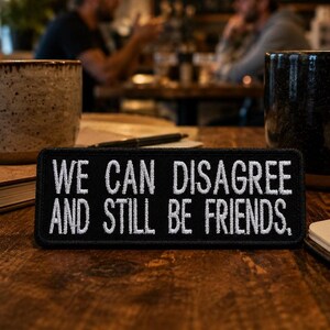 以下が含まれることがあります： 「WE CAN DISAGREE AND STILL BE FRIENDS」と白い文字で書かれた黒い長方形のパッチ。パッチは木製のテーブルの上にあり、コーヒーマグ、ノート、ペンが背景にあります。