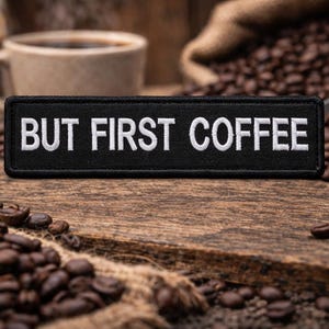 Könnte beinhalten: Ein schwarzer Aufnäher mit weiß gesticktem Text "BUT FIRST COFFEE" liegt auf einer Holzoberfläche, umgeben von Kaffeebohnen. Eine Kaffeetasse und ein Sack voller Bohnen sind im Hintergrund.