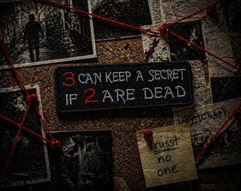 May include: A black patch with the text "3 CAN KEEP A SECRET IF 2 ARE DEAD" in white and red lettering. The patch is on a corkboard with photos, red string, and a note that says "trust no one".