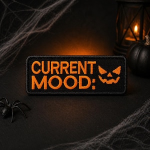 Könnte beinhalten: Schwarzer rechteckiger Aufnäher mit orangefarbenem gesticktem Text "CURRENT MOOD:" und einem Kürbisgesicht. Der Aufnäher befindet sich auf einer schwarzen Oberfläche mit einer Spinne, Spinnweben, einer Laterne und einem Kürbis.