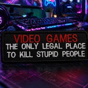 Könnte beinhalten: Schwarzer rechteckiger Aufnäher mit den roten Worten "VIDEO GAMES" und den weißen Worten "THE ONLY LEGAL PLACE TO KILL STUPID PEOPLE". Der Aufnäher liegt auf einem Schreibtisch mit Tastatur und Maus im Hintergrund.