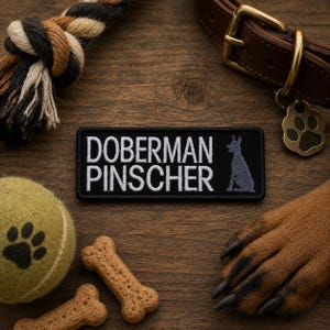 May include: A black patch with white embroidered text reading "Doberman Pinscher" and a silhouette of a Doberman. The patch is surrounded by dog-related items, including a tennis ball, dog biscuits, and a leather collar.