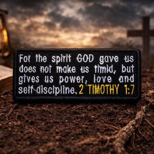 May include: A black embroidered patch with white text quoting 2 Timothy 1:7: "For the spirit GOD gave us does not make us timid, but gives us power, love and self-discipline." A lantern, open book, and cross are in the background.