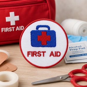 May include: A round, white embroidered patch with a red border and the words "FIRST AID" in red. The patch features a blue first aid kit with a red cross in the center. Other first aid supplies are in the background.