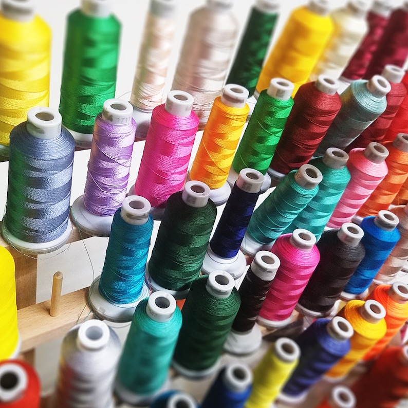 May include: A collection of spools of colorful embroidery floss, arranged on a wooden rack. The floss is organized by color, with shades of blue, green, red, yellow, and purple.