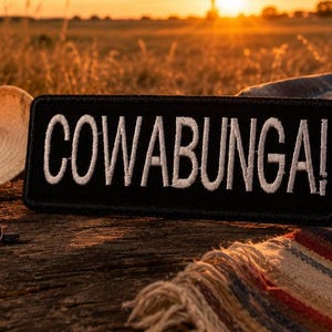 May include: A black rectangular patch with the word "COWABUNGA!" in white, near a straw hat, sunglasses, and a denim jacket. The patch is on a wooden surface with a colorful blanket, with a sunset in the background.