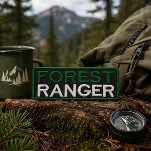Puede incluir: Un parche rectangular con las palabras "FOREST RANGER" en letras blancas y verdes. El parche tiene un fondo oscuro con un borde verde. También se ven una taza esmaltada verde con un diseño de montaña, una brújula y una mochila.