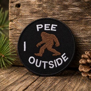 Puede incluir: Un parche redondo negro con texto bordado en blanco y una silueta marrón de Bigfoot. El texto dice "PEE I OUTSIDE". El parche se muestra sobre una superficie de madera, con una rama de pino y una piña en el fondo.