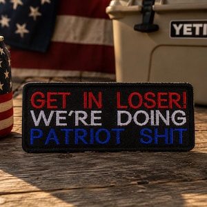 May include: Black rectangular patch with the text "GET IN LOSER! WE'RE DOING PATRIOT SHIT" in red, white, and blue. The patch is on a wooden surface, with an American flag and a can with an American flag design in the background.