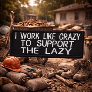 May include: A black rectangular patch with white embroidered text that reads "I WORK LIKE CRAZY TO SUPPORT THE LAZY." The patch is displayed with work boots, a hard hat, and tools, suggesting a construction or labor theme.