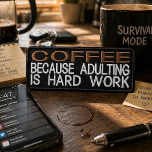Puede incluir: Una taza de café negra con "SURVIVAL MODE" impresa junto a un parche que dice "COFFEE BECAUSE ADULTING IS HARD WORK". También se ven un bloc de notas con una lista de tareas y un teléfono inteligente.