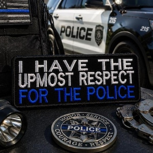 May include: Black patch with white embroidered text: "I HAVE THE UPMOST RESPECT FOR THE POLICE" with blue lettering. A police badge, handcuffs, and a flashlight are also visible. A police car is in the background.