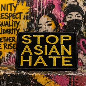 May include: A black rectangular patch with yellow embroidered text that reads "STOP ASIAN HATE." The patch is set against a colorful graffiti background with the words "UNITY, RESPECT, EQUALITY, SOLIDARITY, TOGETHER WE RISE."