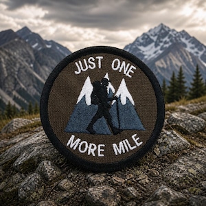 May include: Round patch with the text "JUST ONE MORE MILE" in white, above a silhouette of a hiker with a backpack and trekking pole, and two white mountain peaks with blue shadows. The patch has a brown background and a black border.