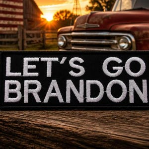 May include: Black rectangular patch with white embroidered text reading "LET'S GO BRANDON." The patch is on a wooden surface, with an American flag, a red vintage truck, and a cap in the background.