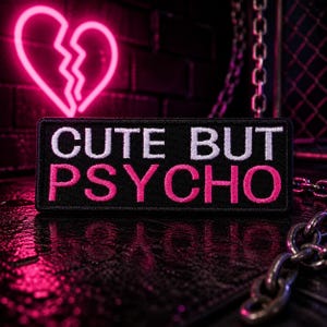 May include: A black rectangular patch with the words "CUTE BUT PSYCHO" embroidered in white and pink. A neon pink broken heart is in the background. The image has a dark, edgy aesthetic with a chain in the foreground.