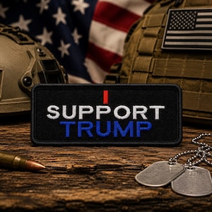 May include: A black embroidered patch with the words "SUPPORT TRUMP" in white and blue, with a red stripe. The patch is on a wooden surface, with a military helmet, American flag, and dog tags in the background.