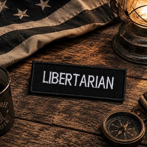 May include: A black patch with the word "LIBERTARIAN" embroidered in white. The patch is on a wooden surface, alongside a vintage lantern, a compass, a mug, and a distressed American flag.