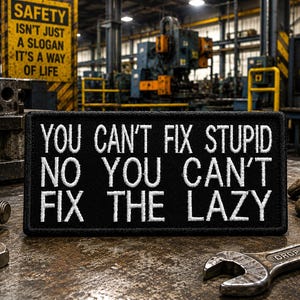 May include: A black rectangular patch with white embroidered text that reads "YOU CAN'T FIX STUPID NO YOU CAN'T FIX THE LAZY." The patch is on a metal surface with tools and a sign that says "SAFETY ISN'T JUST A SLOGAN IT'S A WAY OF LIFE."