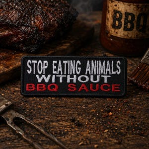 May include: A black patch with white and red embroidered text that reads "STOP EATING ANIMALS WITHOUT BBQ SAUCE." The patch is on a wooden surface with a BBQ sauce bottle and a piece of cooked meat in the background.
