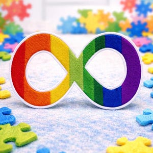 Puede incluir: Un parche bordado con el símbolo del infinito en colores del arcoíris, con un borde blanco, es el elemento central. La imagen está salpicada de coloridas piezas de rompecabezas. El fondo es azul claro, creando una imagen brillante y simbólica.