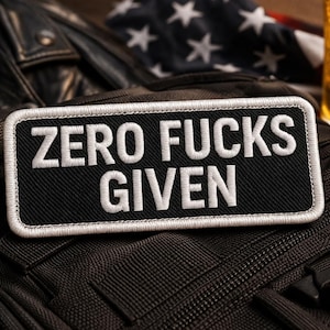 May include: Black rectangular patch with white embroidered text reading "ZERO FUCKS GIVEN." The patch is bordered with white stitching. The patch is on a black tactical backpack. A glass of beer with a skull and crossbones is in the background.