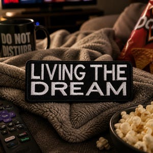 May include: A black patch with white embroidered text that reads "LIVING THE DREAM" is displayed on a gray blanket. A black mug with "DO NOT DISTURB" is visible, along with a bowl of popcorn and a remote control.
