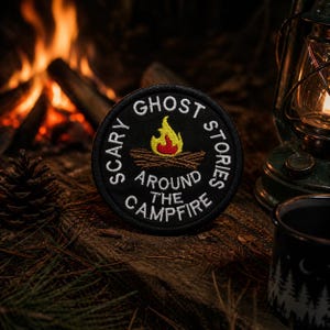 May include: A circular black patch embroidered with the words "Scary Ghost Stories Around the Campfire" in white. The patch features a campfire graphic with a yellow and red flame. The patch is designed for outdoor enthusiasts.