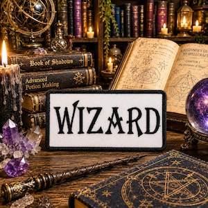 May include: A patch with the word "WIZARD" in black letters on a white background. The patch is surrounded by a black border. The patch is on a wooden table with books, candles, crystals, and a magic wand.