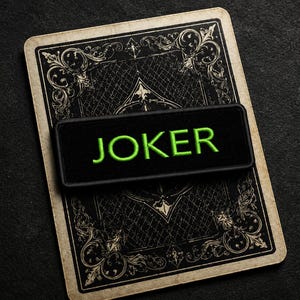 May include: A black patch with the word "JOKER" embroidered in green, placed on a playing card. The card's back features an ornate, vintage design. The background is a dark, textured surface.