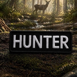 Puede incluir: Un parche rectangular negro con la palabra "HUNTER" bordada en blanco. El parche se encuentra sobre un fondo de bosque con un ciervo a lo lejos, lo que sugiere un tema de caza o actividades al aire libre.