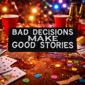 Puede incluir: Un parche negro con texto bordado en blanco que dice "BAD DECISIONS MAKE GOOD STORIES". El parche está sobre una superficie de madera con líquido derramado, naipes y vasos de plástico rojos. Botellas de cerveza y luces de colores en el fondo.