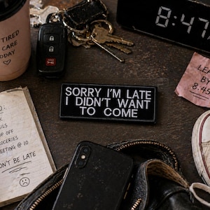 May include: A still-life composition featuring everyday items. A coffee cup with text, a to-do list, a phone, keys, a patch with text, a clock, and a sneaker. The patch reads "SORRY I'M LATE I DIDN'T WANT TO COME".