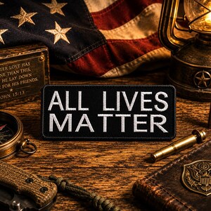 May include: A black patch with the words "ALL LIVES MATTER" in white, alongside an American flag, a vintage lantern, and a compass. Other items include a knife, a small box with text, and a leather-bound notebook.