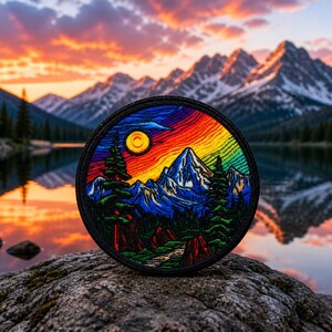 May include: A round embroidered patch featuring a mountain landscape with a sunset sky. The patch has a black border and depicts a mountain range, trees, and a lake. The sky transitions from orange to blue, with a yellow sun.