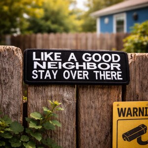 Può includere: Una toppa rettangolare nera con testo bianco che recita "LIKE A GOOD NEIGHBOR STAY OVER THERE" è attaccata a una staccionata di legno. La toppa è centrata sulla recinzione, con uno sfondo sfocato di alberi e una casa blu.