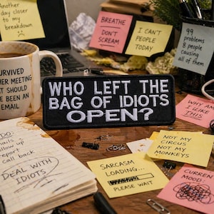 May include: A desk scene with a coffee mug, notepad, and sticky notes. The mug reads "I survived another meeting that should have been an email." A patch says "WHO LEFT THE BAG OF IDIOTS OPEN?" Other notes have humorous phrases.