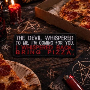 May include: A black embroidered patch with the text "THE DEVIL WHISPERED TO ME, I'M COMING FOR YOU, I WHISPERED BACK, BRING PIZZA." is displayed on a wooden surface. Pizza slices and a lit candle are also visible.