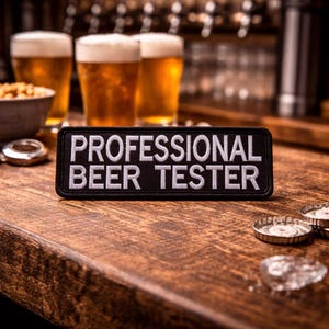 May include: A black patch with white text that reads "PROFESSIONAL BEER TESTER" sits on a wooden bar. Three glasses of beer with foamy heads are in the background, along with a bowl of snacks and bottle caps.